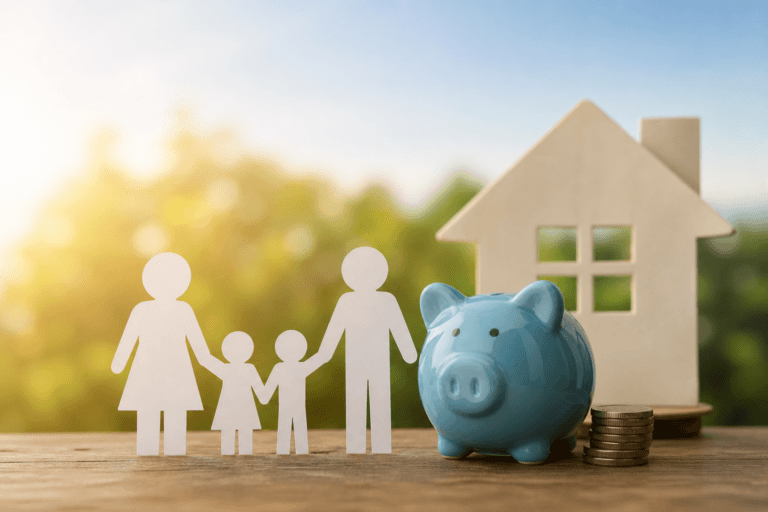 Paper cutout figures of a family standing beside a blue piggy bank, stacked coins, and a small house model on a wooden surface, symbolizing financial support and stability for working families.